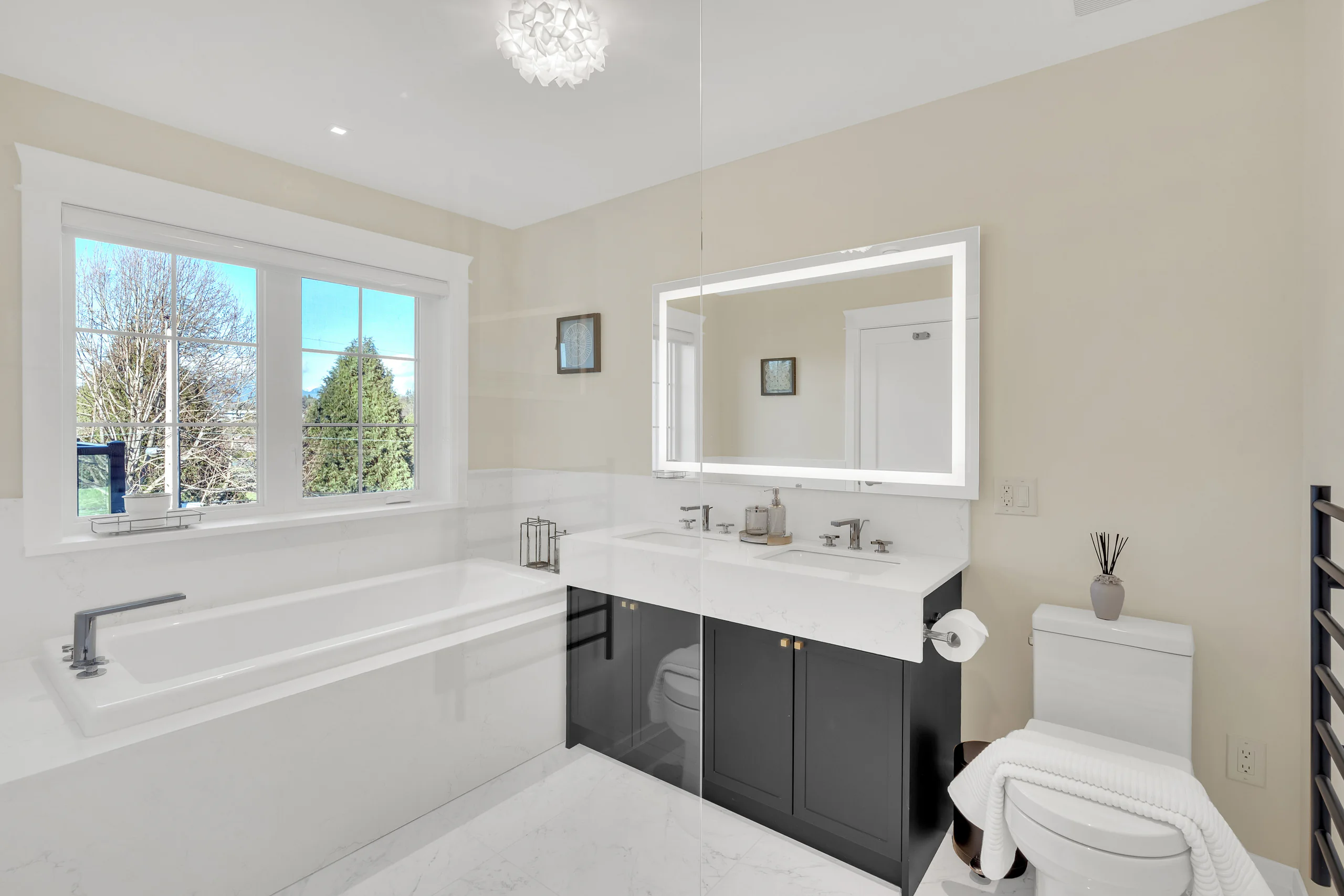 A white bathroom with a lit mirror, a dark vanity and a window
