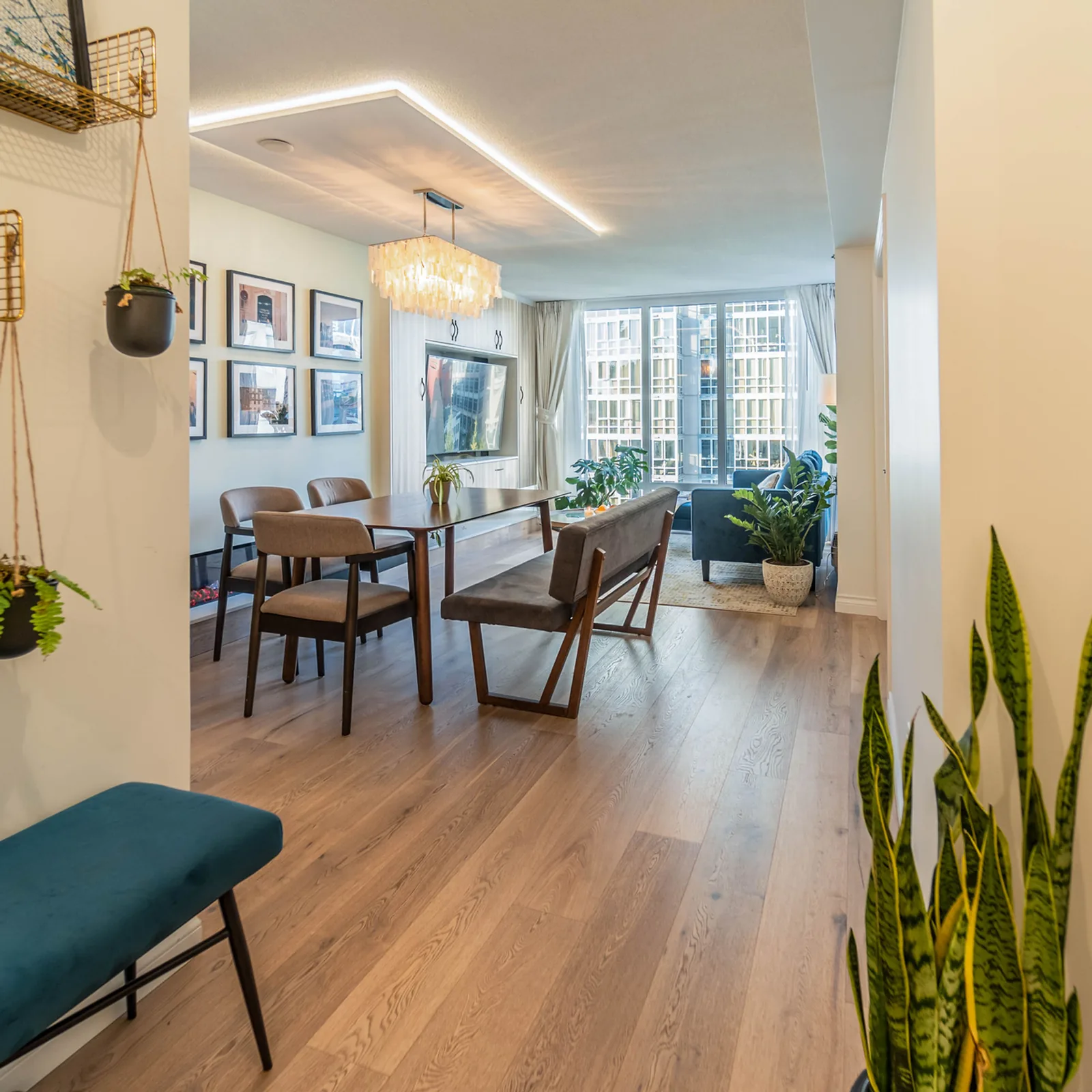 Open living and dining room in a condo with wide-plank wood floors, a long dining table with a bench, a lit ceiling panel and floor-to-ceiling windows